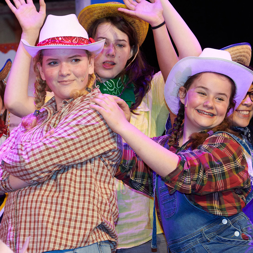 Four actresses on stage dressed as farm hands in cowboy hats, overalls, jeans and scarfs.